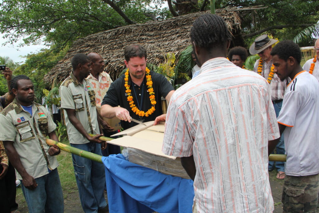Um homem usando um colar de flores alaranjadas usa uma faca para abrir uma caixa segurada por pessoas em postes de madeira enquanto várias pessoas observam.