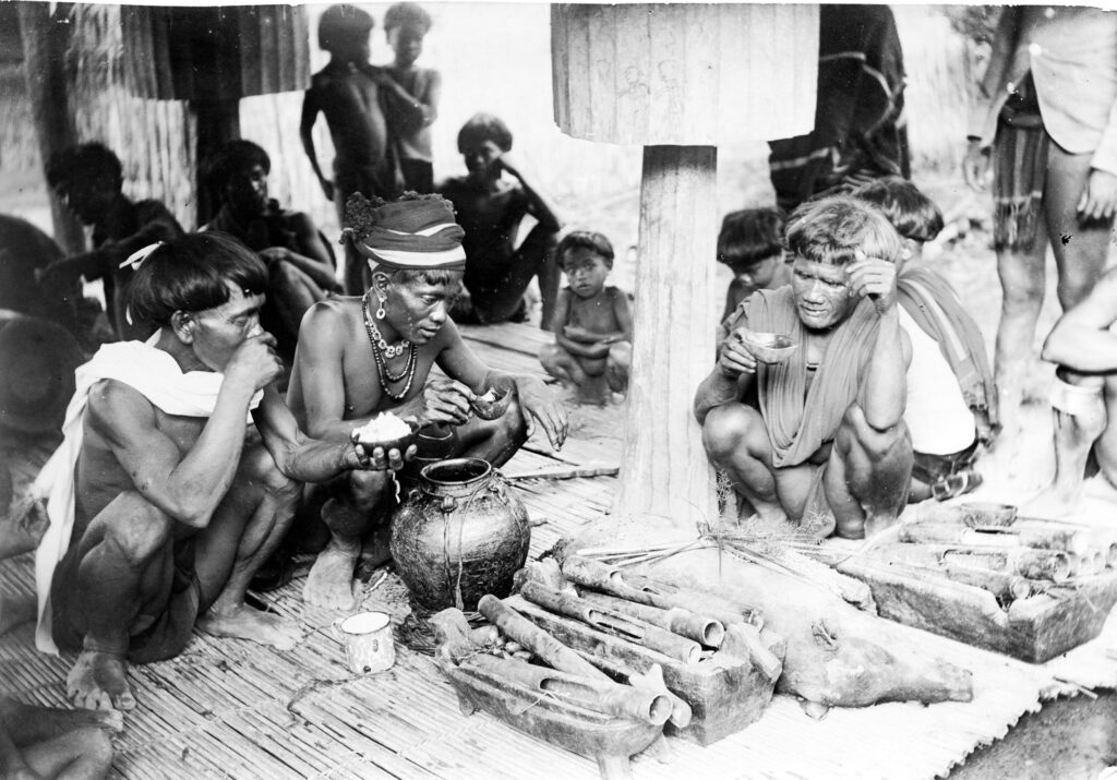 A black-and-white image features three people wearing scarves and beaded necklaces squatting near a large black pottery vase and objects lying in wooden containers. Behind them, other adults and children gather.