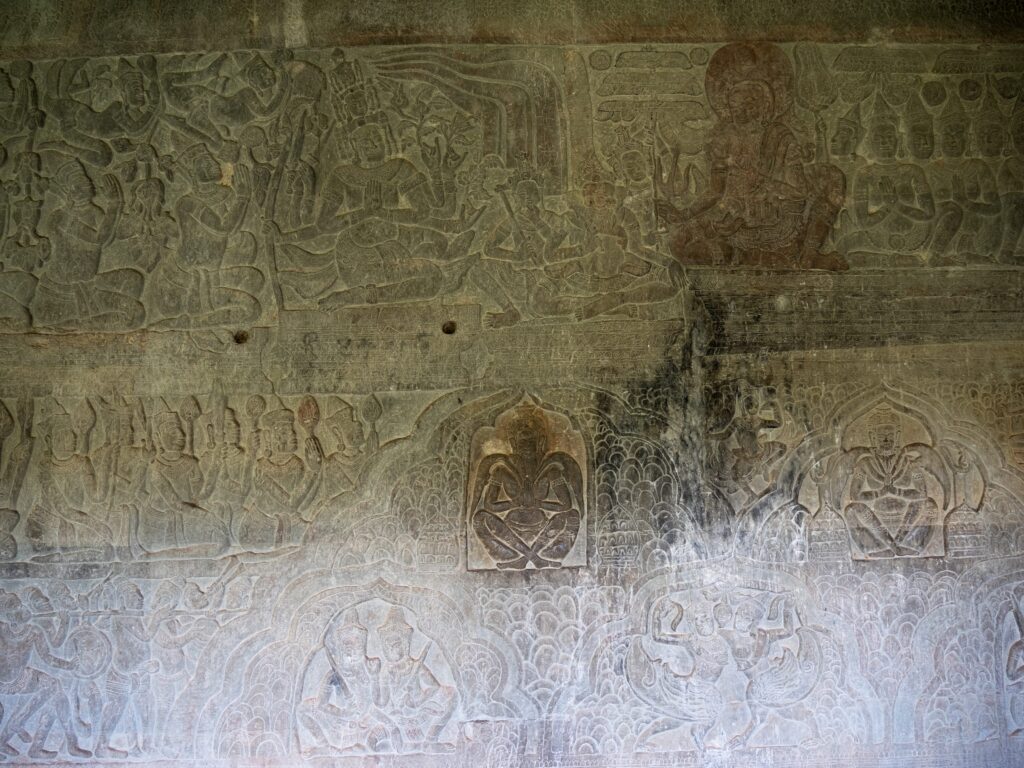 A tan wall features an engraved image of a person sitting cross-legged and holding their hands together in front of their chest. Surrounding this image are many additional engravings of people doing a variety of activities.