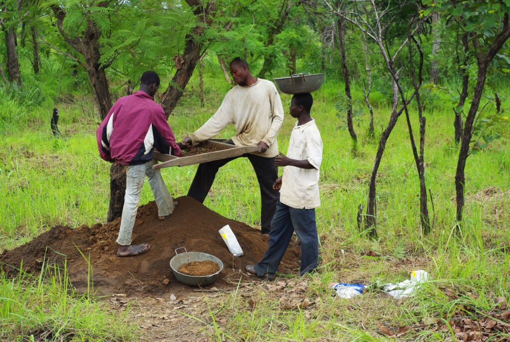 Three people stand atop a mound of earth surrounded by green grass and small trees. Two of them hold a tool to sift soil while the third balances a bowl on his head.