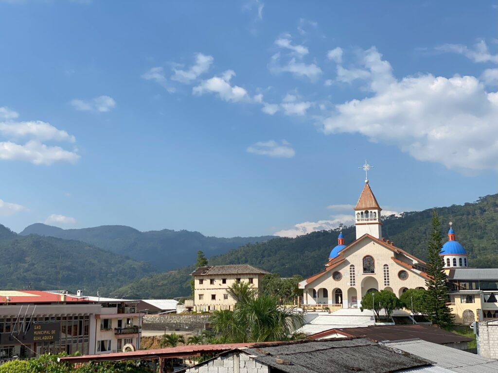 A scenic view of a town features a prominent church with red and blue domes under a clear sky and set against a backdrop of lush, green mountains.