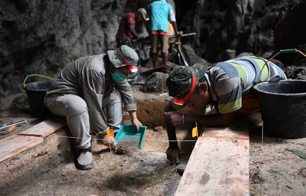 In the foreground, two people with headlamps use tools to dig into the earth, surrounded by buckets, hand tools, and two small yellow flags marking off the section they work in. In the unfocused background, several people stand at a screen looking for ancient objects.