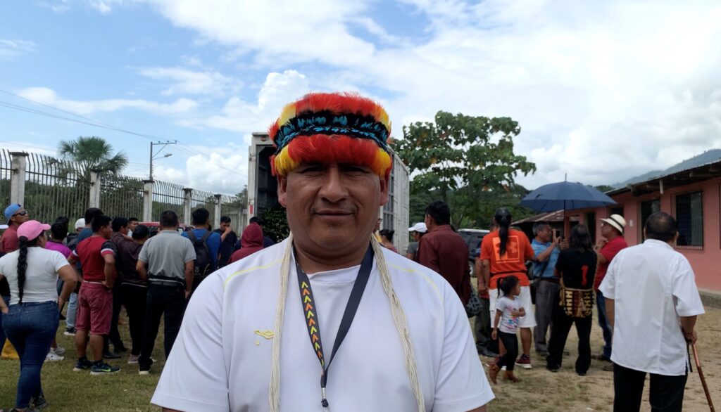 A man wearing an orange, yellow, black, and blue feathered headband stands amid a gathering of people in an outdoor setting with a blue sky overhead.