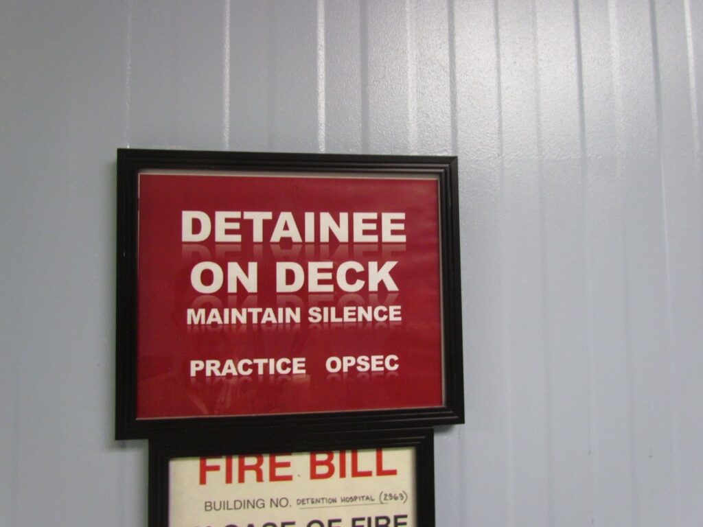 A black-bordered sign reads "Detainee on Deck, Maintain Silence, Practice OPSEC."