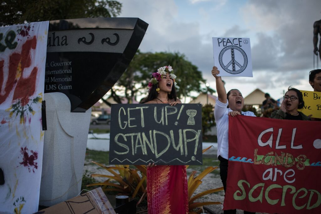 A group of people stands outside holding signs displaying various pro-peace messages such as “Get Up! Stand Up!” and “All Lands Are Sacred.”
