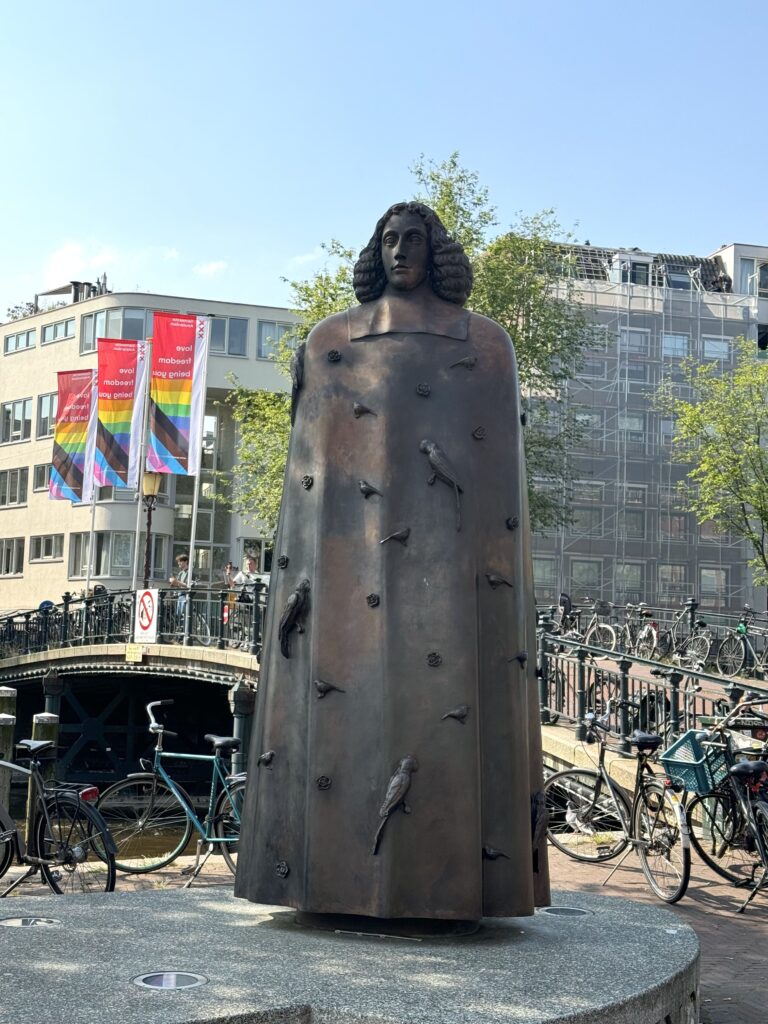 In front of a foot bridge lined with parked bicycles, a dark metallic statue of a person stands on a gray stone pedestal.