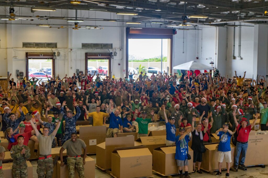 In a warehouse, hundreds of people wearing red, green, and blue T-shirts and army fatigues stand among cardboard boxes with their arms aloft.