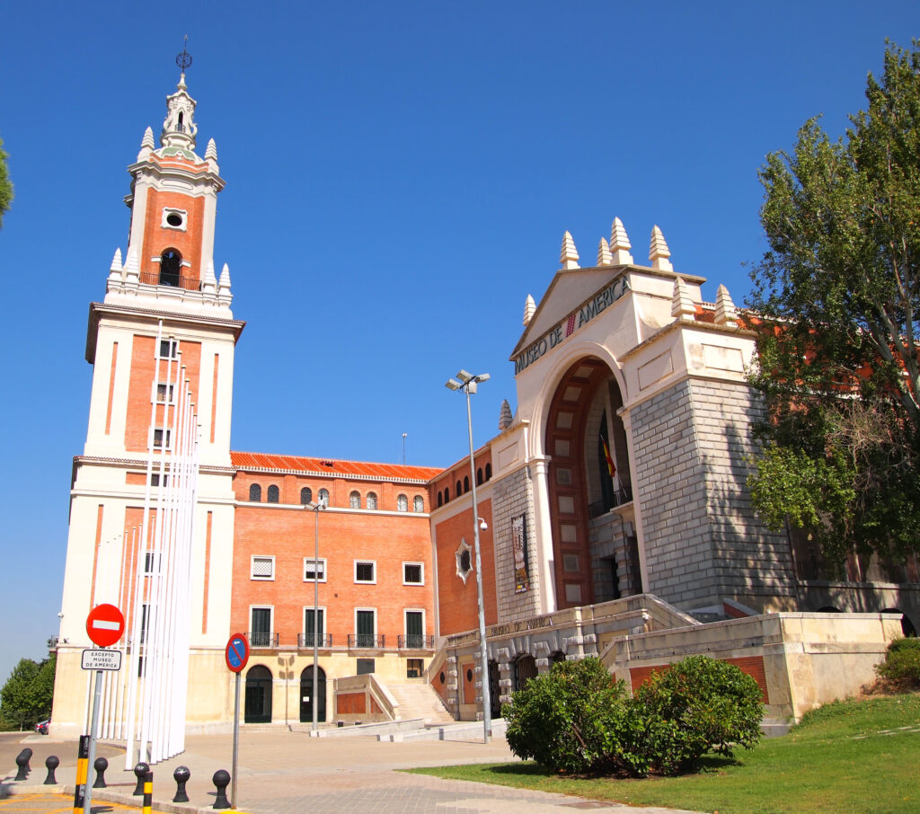 Under a cloudless blue sky and next to a green lawn with a tree and a low bush stands an expansive building of red and gray bricks and white stucco.