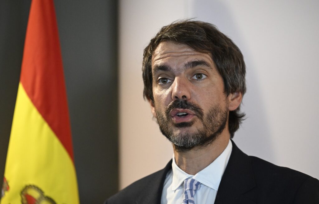 A bearded man wearing a dark suit and a tie speaks next to a red and yellow flag.
