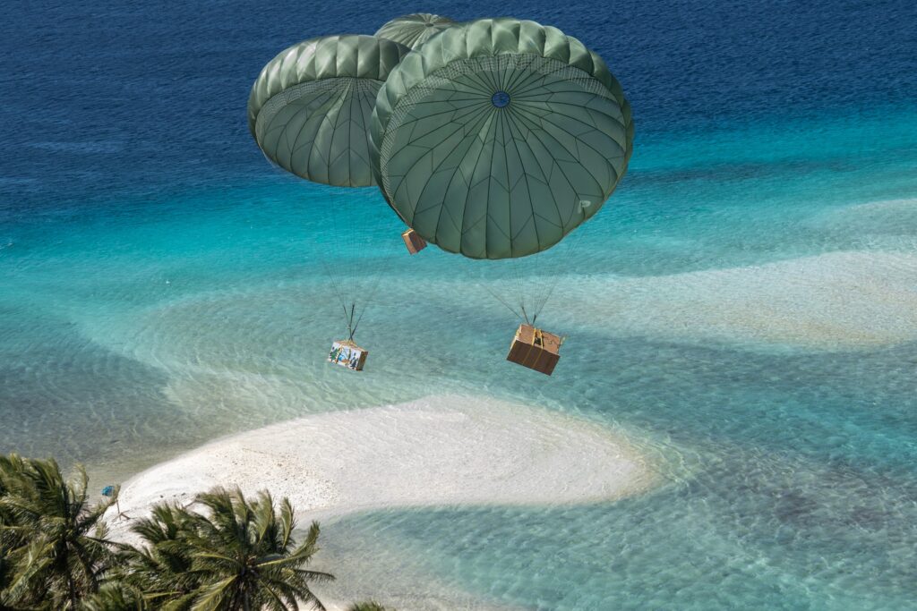 Boxes attached to teal-green parachutes float through the air above turquoise blue waters, a small stretch of white-sand beach, and palm trees.