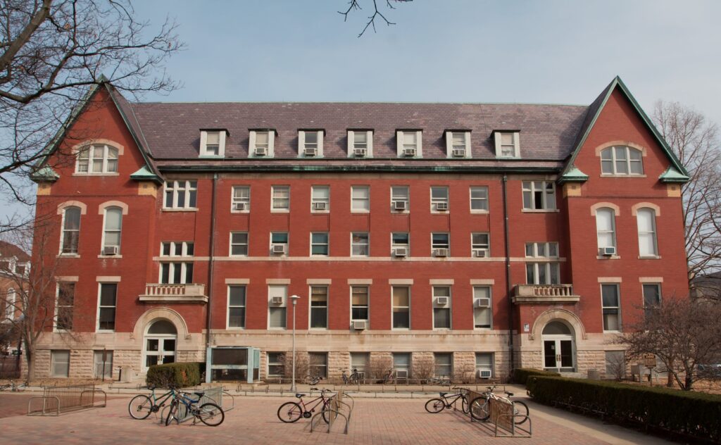 A few bicycles stand locked to racks in front of the broad façade of a red brick Victorian building.