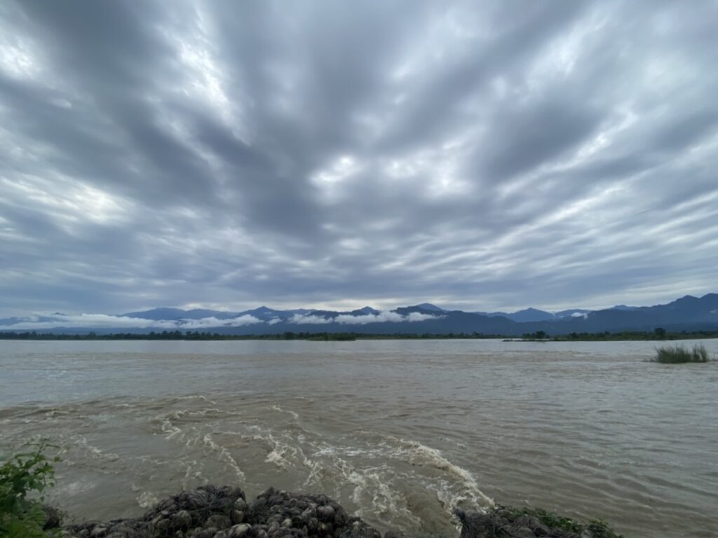 Gray clouds drift above a wide, brown river. Mountains loom on the horizon.