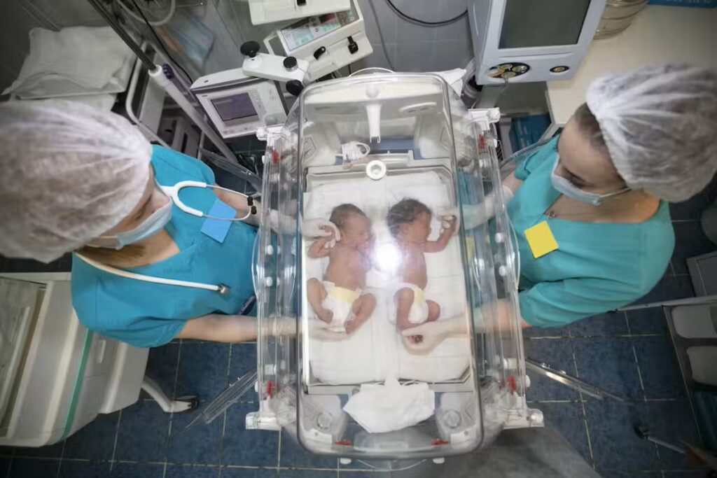 Newborn twins lie side by side in a neonatal incubator, a temperature-controlled small hospital bed with a hard, clear plastic cover. They are monitored by two medical staff wearing surgical caps, masks, and blue-green scrubs.