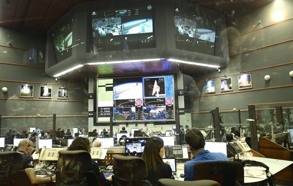 In a large room with high ceilings, people sitting at desks monitor several large screens that display video feeds and technical information.
