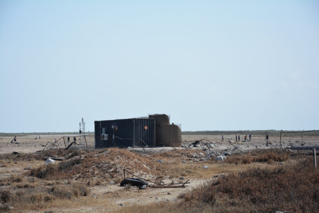 A black corrugated metal container sits in the center of a flat, brownish patch of ground where twisted hunks of metal fencing and other debris are scattered. In the background, a few people slowly walk across a field.