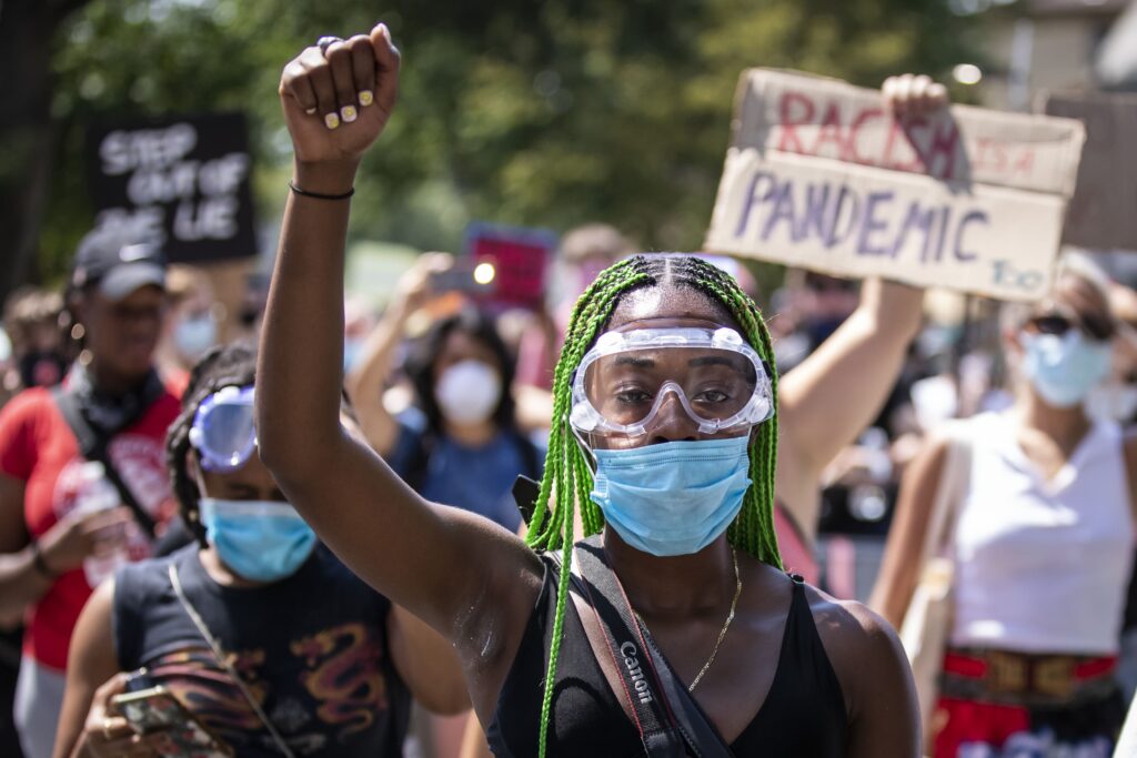 A person with long, green, braided hair, goggles, and a surgical mask holds up their fist in a gesture of protest. Behind them, among a crowd of other demonstrators, someone holds a sign that reads: “Racism is a pandemic too.”