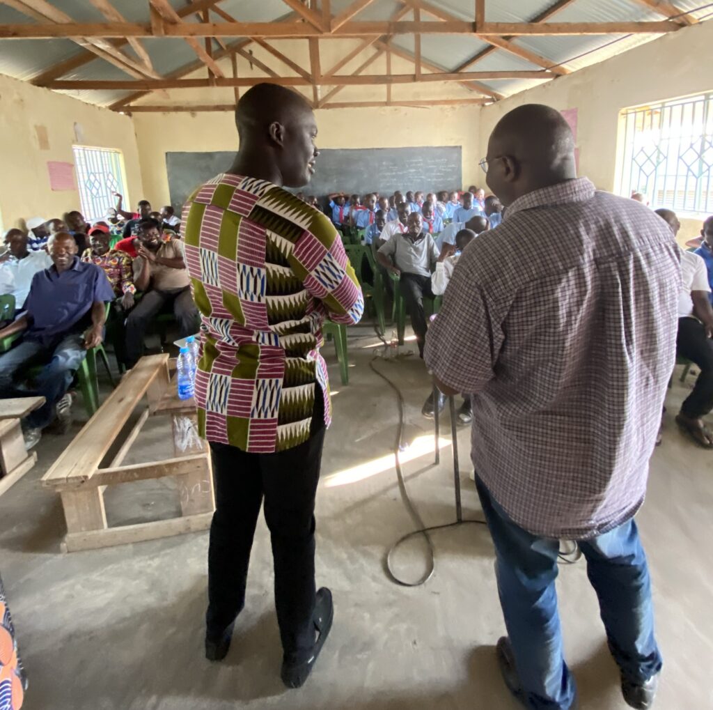 Two people stand before a full classroom with a corrugated metal ceiling and exposed wooden beams above.