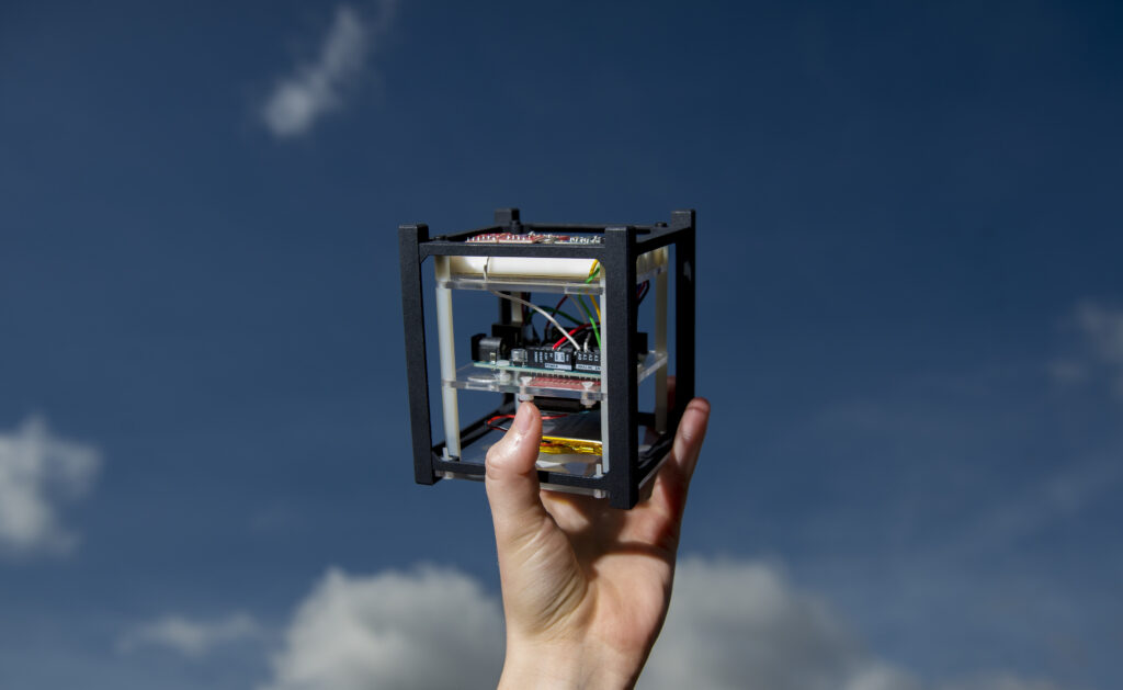 Against a blue sky dotted with fluffy white clouds, a hand holds aloft a hollow cube filled with white, black, red, green, and yellow electronic components.