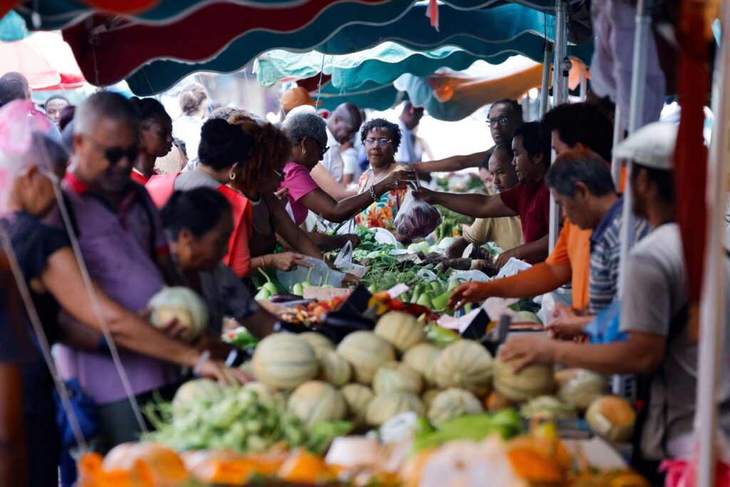Customers and vendors interact and do business across tables of colorful fruit and other produce. Green, orange, blue, and pink colors predominate.