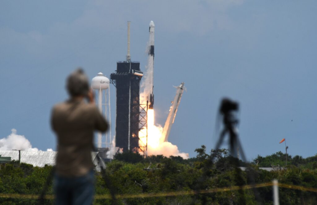 A blurry figure photographs a large white rocket as it lifts off its launch pad and scaffolding, emitting a fiery plume of exhaust.