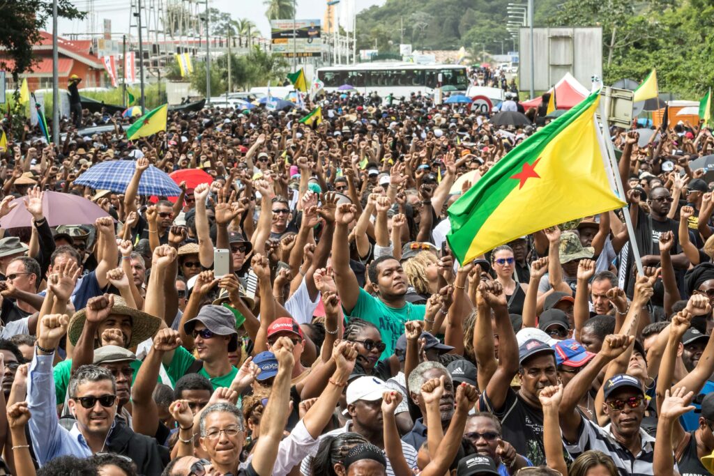 A large crowd holds its fists aloft in unison. Some members of the crowd hold up green and yellow flags with a red star in the middle and umbrellas.