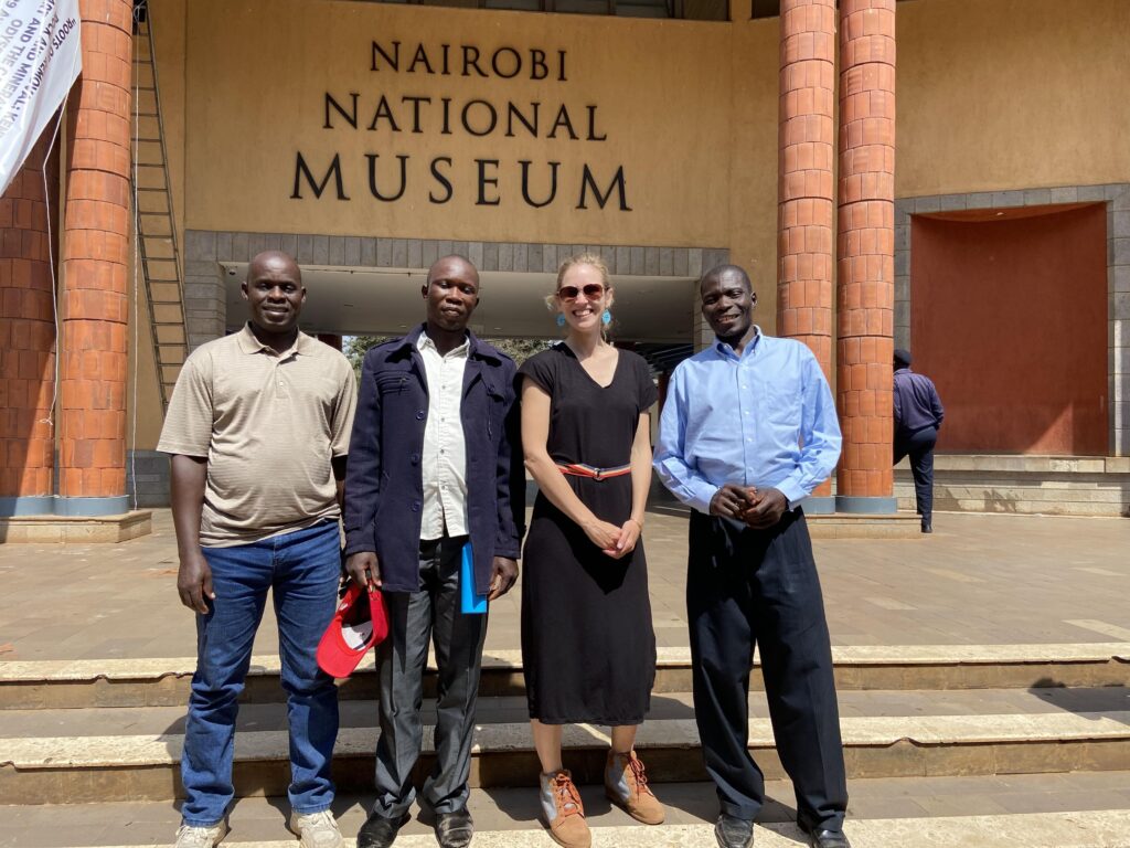 Four people stand in front of the entrance to a building whose façade is tan and is framed by large red columns.