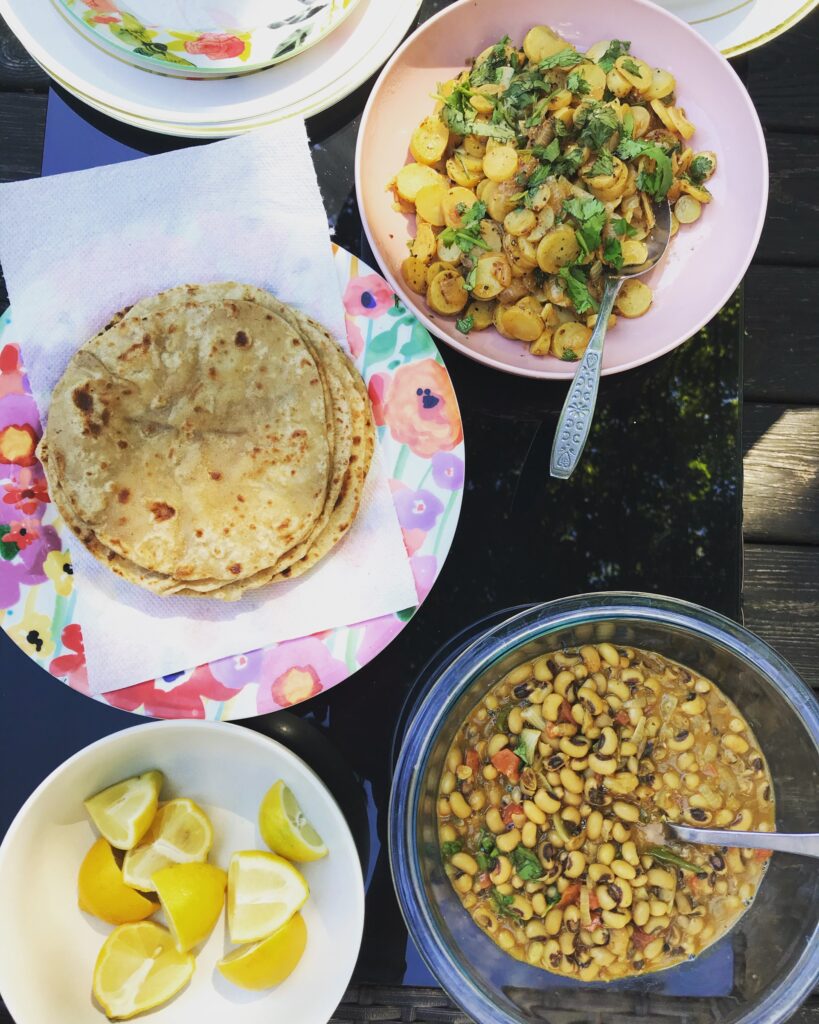 A colorful spread of food dishes includes a stack of flatbread, potato curry garnished with herbs, a bowl of light-colored beans, and a plate of sliced yellow lemons.