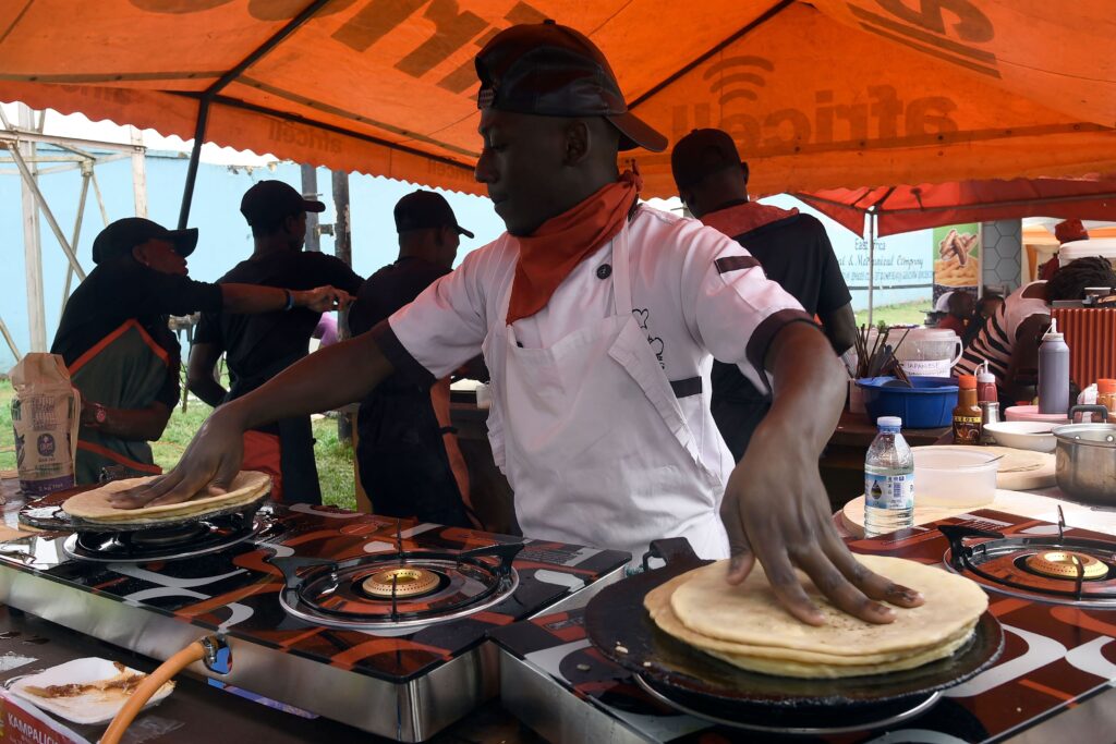 Underneath a large orange tent, a man wearing a backward baseball hat, red handkerchief, and white shirt and apron uses both hands to fry flatbread on multiple burners. Behind him, a small group wearing dark clothes and black baseball hats congregates.