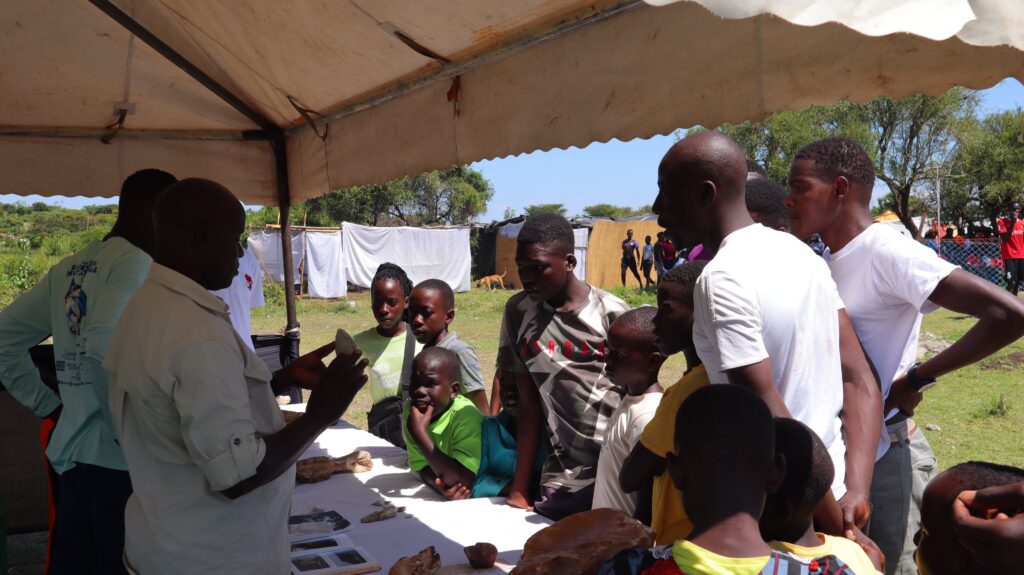 Beneath a large white tent, two people stand at a table explaining a pointed object to a small crowd of interested onlookers—both children and adults.