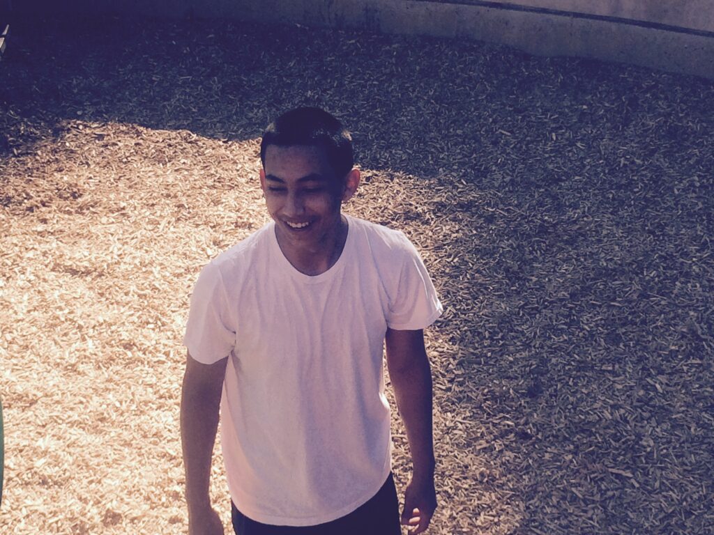 A young man in a white T-shirt smiles while standing in a courtyard.