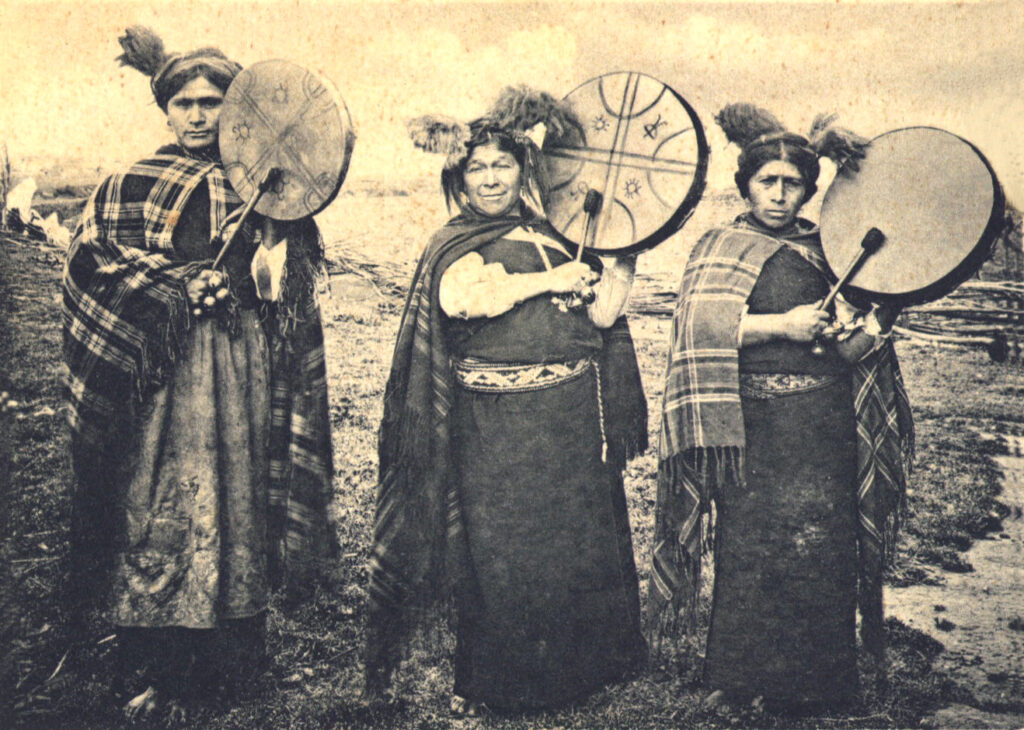 A sepia-toned photograph shows three people dressed in long dark tunics and plaid shawls holding up disc-shaped drums and mallets, which are used to play the drums.