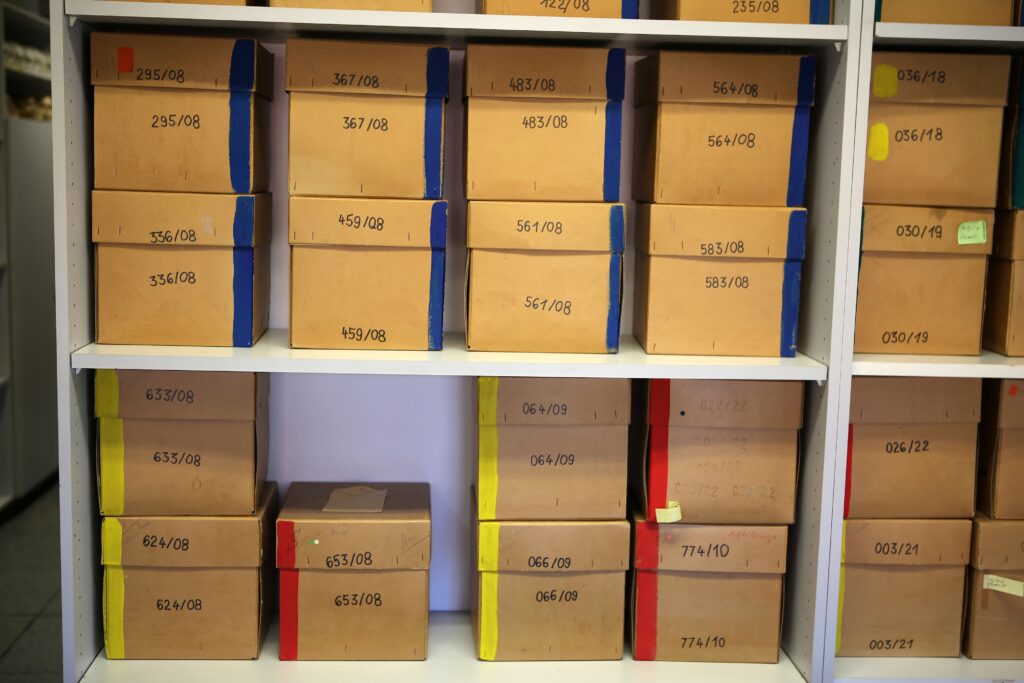 Light brown cardboard boxes color coded with blue, yellow, and red stripes and labeled with black marker are stacked on white shelves.
