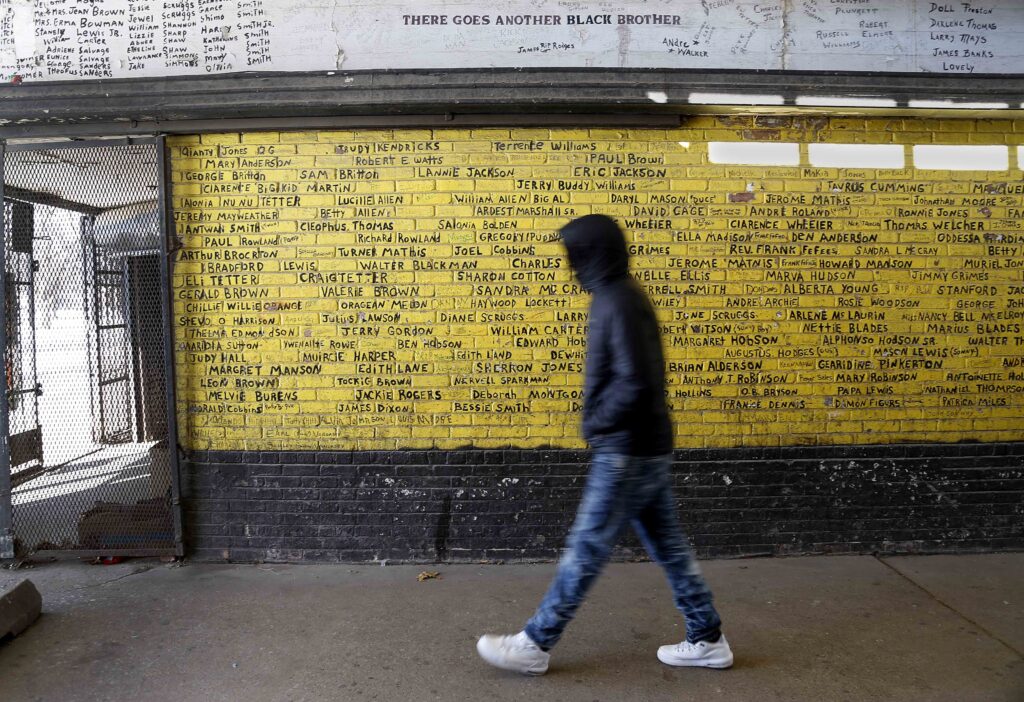 A person in blue jeans, white sneakers, and a hooded sweatshirt walks past a yellow wall with black words painted on it.