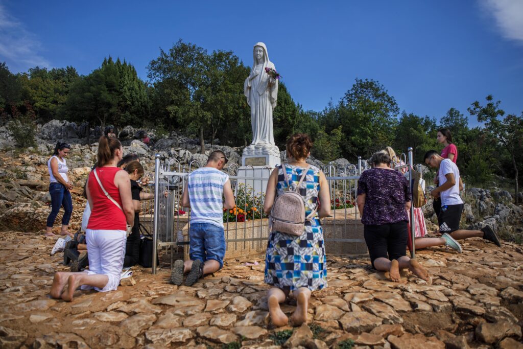 Under a blue sky, a small group of people kneel on rocky beige ground beside a statue of a woman carved from gray stone. Leafy green trees are in the background.