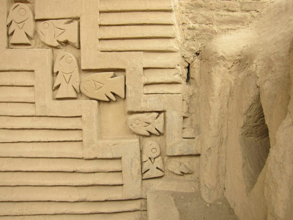 A close-up of a tan-colored wall features deep, lined grooves and carved fish designs in relief within a zigzag frame.