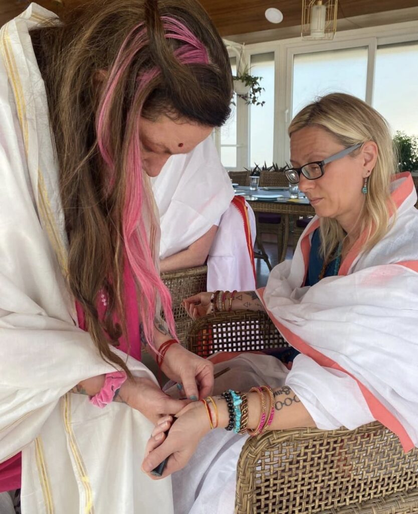 A woman with pink highlights in her long hair wears a white robe and touches the wrist of a second white-robed woman sitting in a wicker chair.