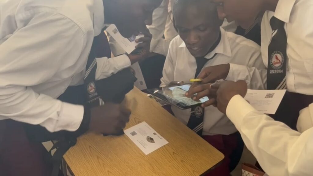 Students wearing white shirts and ties gather excitedly around a computer tablet.