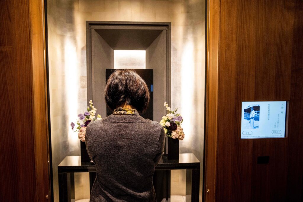 A person bows in a wood-paneled vestibule in front of a black table that holds two small vases of flowers.