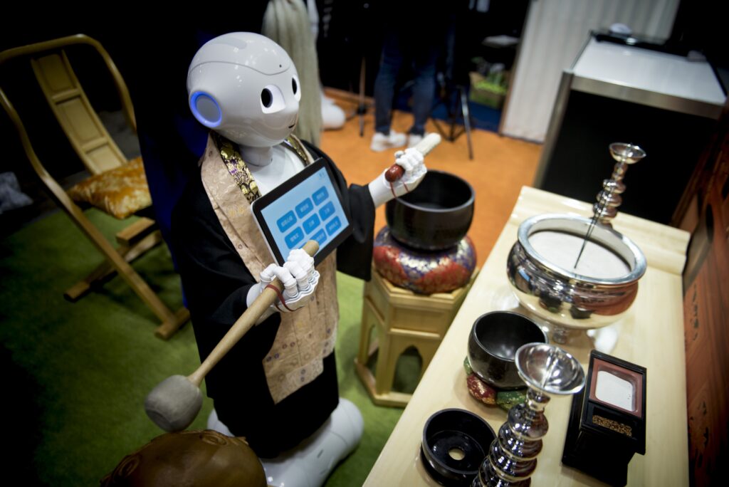 A white robot wearing a kimono and a tablet screen hanging around its neck holds a mallet in its right hand and a white stick with a red end in its left hand. It stands in front of silver bowls and other accoutrement.