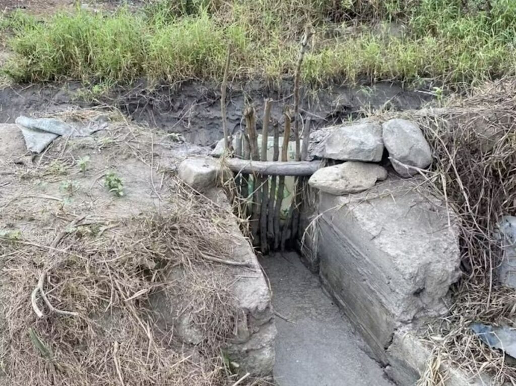 A wooden gate sits between two slabs of cement blocks with scraggles of brush, dirt, and stones on either side.