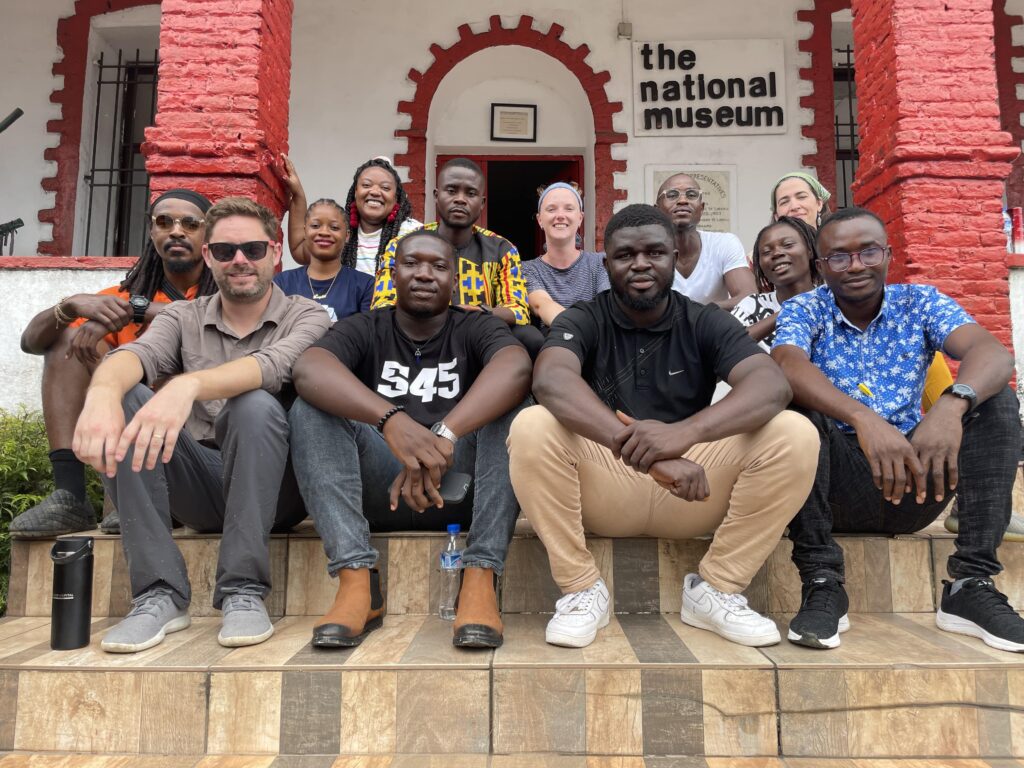 Two rows of people sit on steps outside the entrance of a building with white walls and red columns.