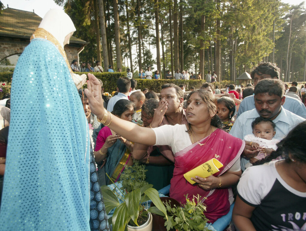 Standing at the front of a crowd of people in a verdant park with bushes and tall trees, a woman in a dark-pink sari reaches out to touch a statue of a woman clad in a turquoise cloak and white hood.