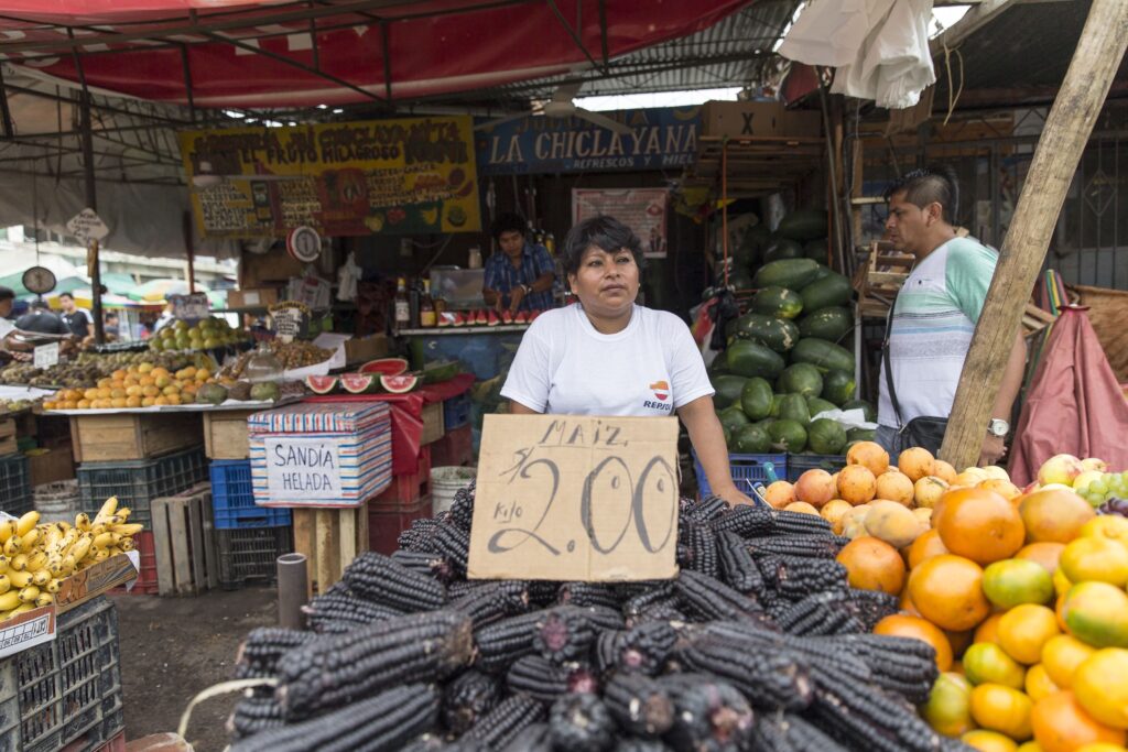 A person with short black hair wearing a white shirt stands behind a table filled with a pile of purple corn on the cob and bright oranges in a farmers’ market. A price sign on brown cardboard sits atop the corn, with other tables filled with fruits and vegetables.