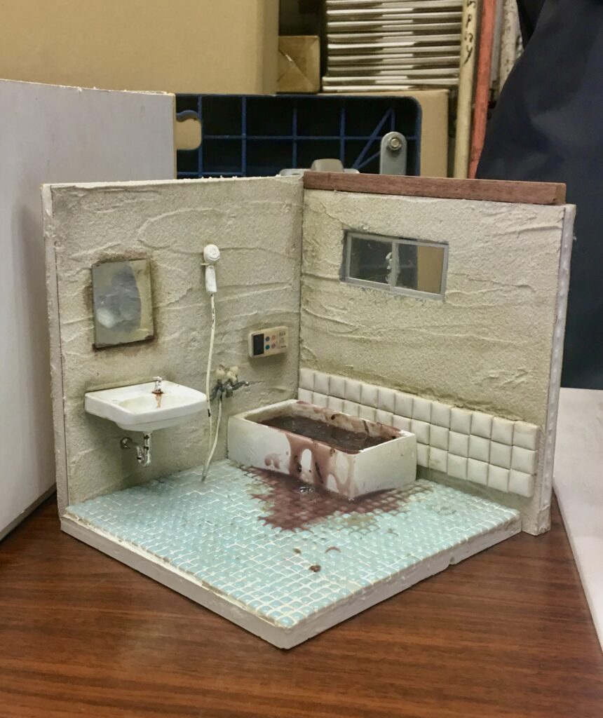 A small diorama depicts a bathroom with a bathtub filled with a dark liquid overflowing on blue tile.