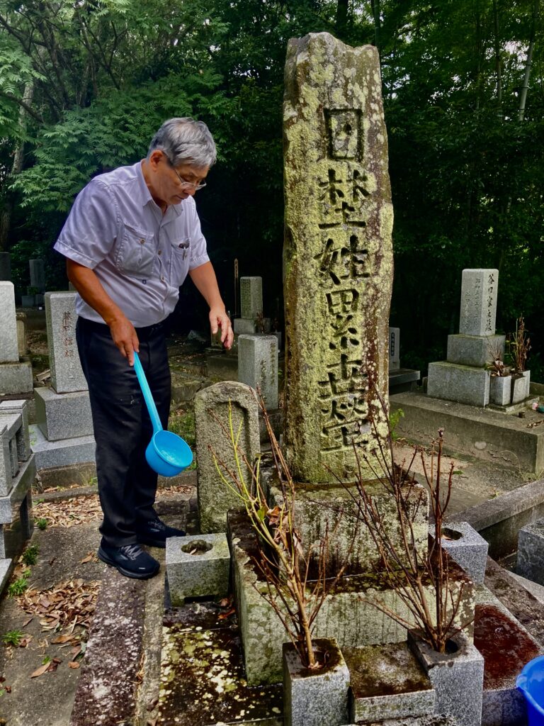 A man holding a long-handled blue plastic vessel stands beside a tall, mossy stone monument in a leafy green cemetery.
