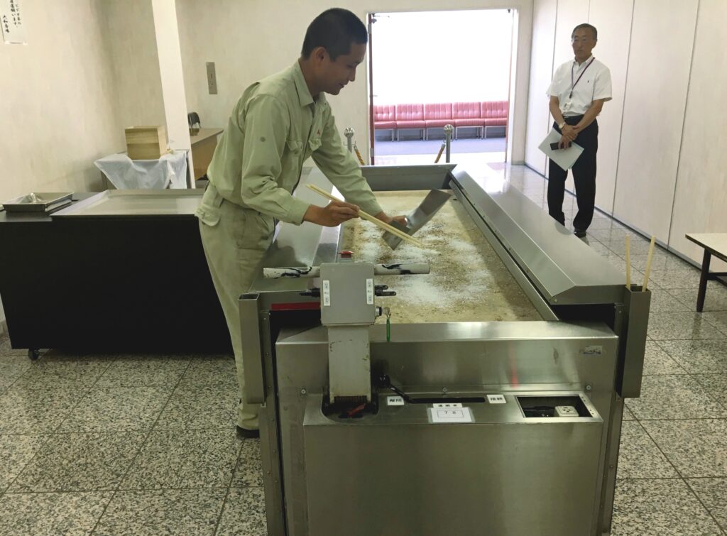Two people stand in a room with a tile floor and white walls. One stands in front of a large metal countertop holding long chopsticks. The other looks on.