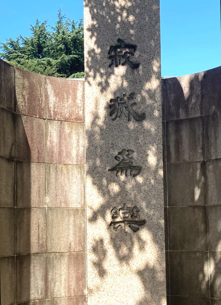 A long, tall stone monument with Japanese characters inscribed on it sits partially enclosed by a cylindrical concrete wall. Behind the wall, the top of a green bush is visible against a clear blue sky.