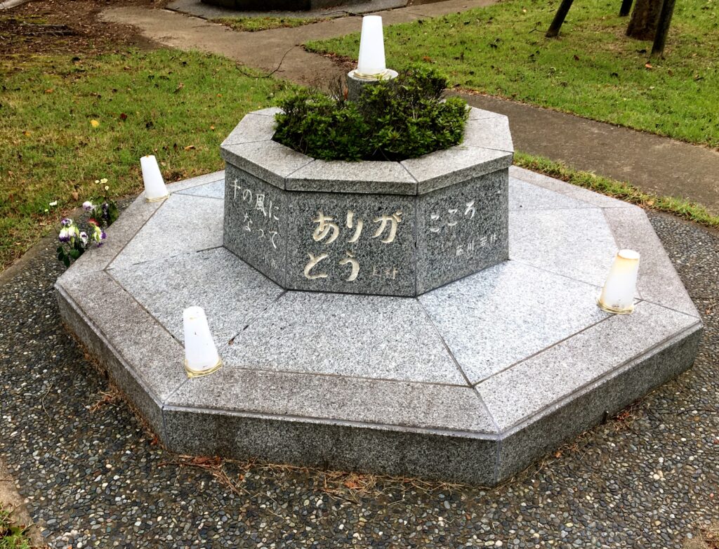 A low-lying, octagonal stone monument is surrounded by gravel and green grass. In the center, a small bush grows. Ann