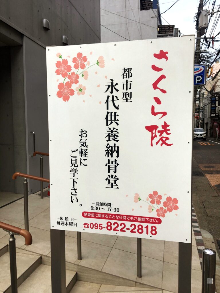 A white sign with red and black Japanese characters and pink flowers stands outside a gray building.