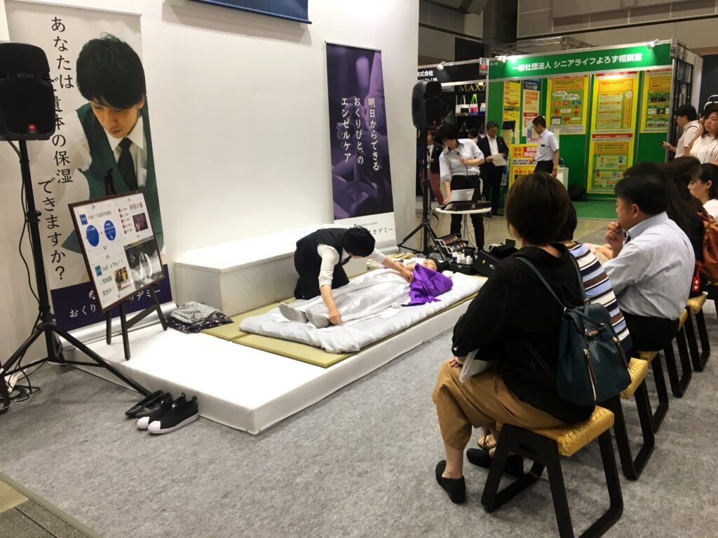 A person on a small stage flanked by black speakers kneels while folding a piece of gray cloth. A row of seated audience members looks on.